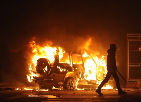 a silhouetted person walks past a burning car in the dark.