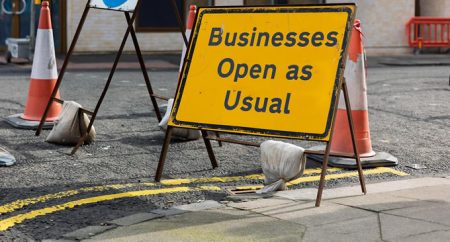 Yellow sign reading 'Businesses Open as Usual' surrounded by traffic cones on a city street.