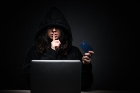 Person in a dark hoodie holding a credit card and making a shushing gesture in front of a laptop.
