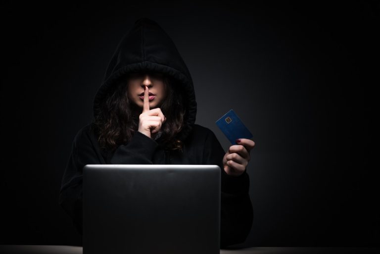 Person in a dark hoodie holding a credit card and making a shushing gesture in front of a laptop.