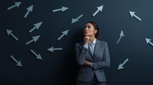 Pensive businesswoman standing against a dark wall with many arrows pointing in different directions, symbolizing a difficult decision making process.