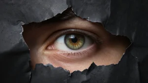Close-up of a green-brown iris peering through a jagged tear in dark paper or wall material.
